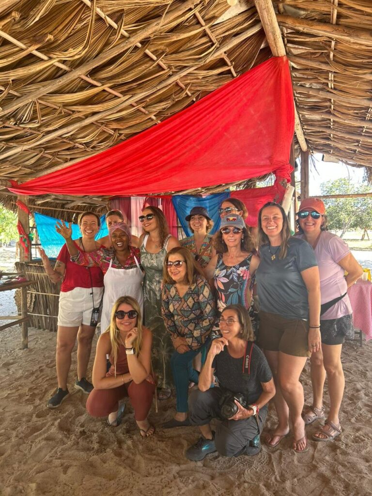 Grupo de mulheres em um ambiente rústico com um telhado de palha e tecidos coloridos.